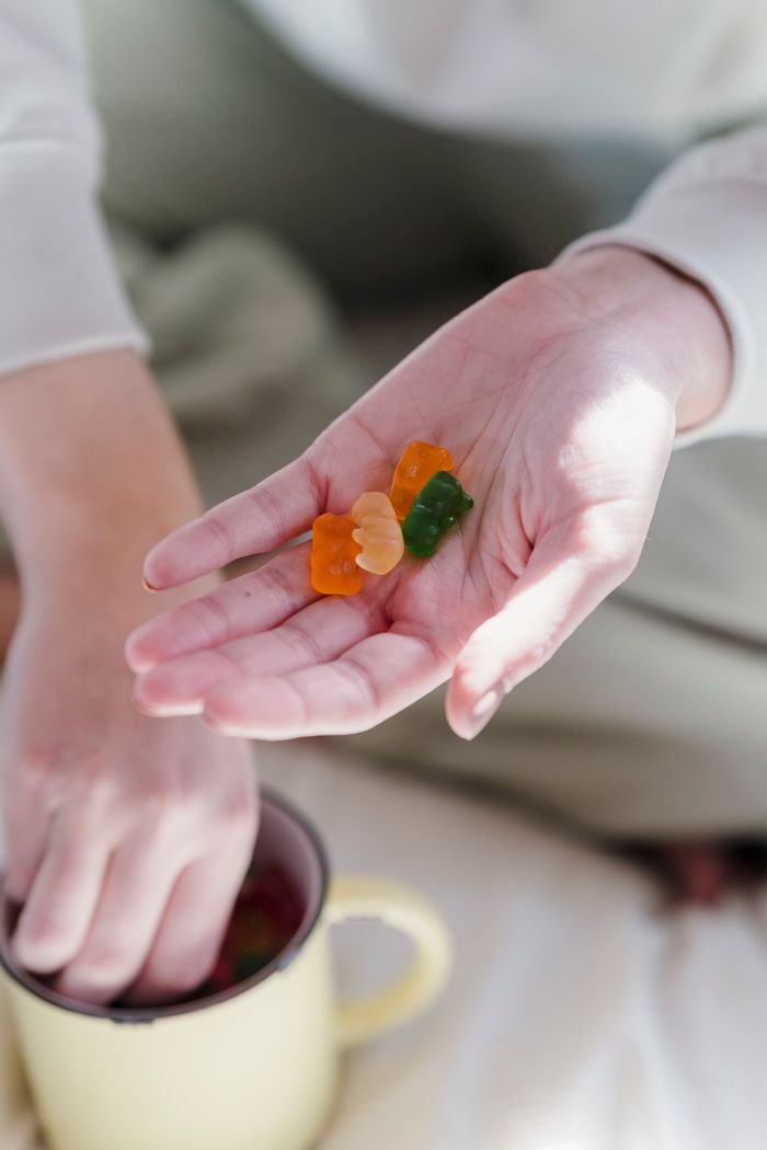 From above of crop anonymous female holding handful of yummy colorful gummy bears and drinking hot coffee on cozy bed